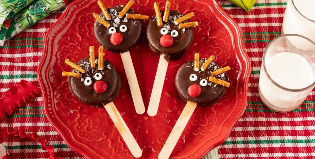 Four chocolate-covered cookies on sticks are decorated as reindeer with pretzel antlers, candy eyes, and red candy noses. They are arranged on a red plate over a festive plaid tablecloth, with glasses of milk nearby.