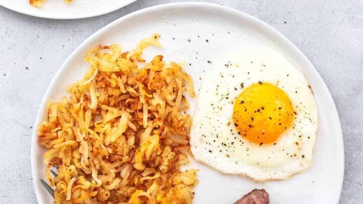 Two plates of breakfast featuring crispy hash browns, a sunny-side-up egg, and two sausages. The plates are placed on a light gray surface, and each has a fork resting on the rim.