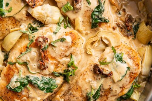 A skillet filled with creamy Tuscan chicken, featuring pan-seared chicken breasts smothered in a rich, creamy sauce with sun-dried tomatoes, spinach, and artichokes. The dish is garnished with fresh herbs.