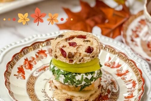A fall-themed plate with a stack of turkey tea sandwiches, garnished with herbs. Decorated with autumn leaves and a small pumpkin illustration, the plate features an elegant floral pattern. A cup with a floral design sits in the top corner.