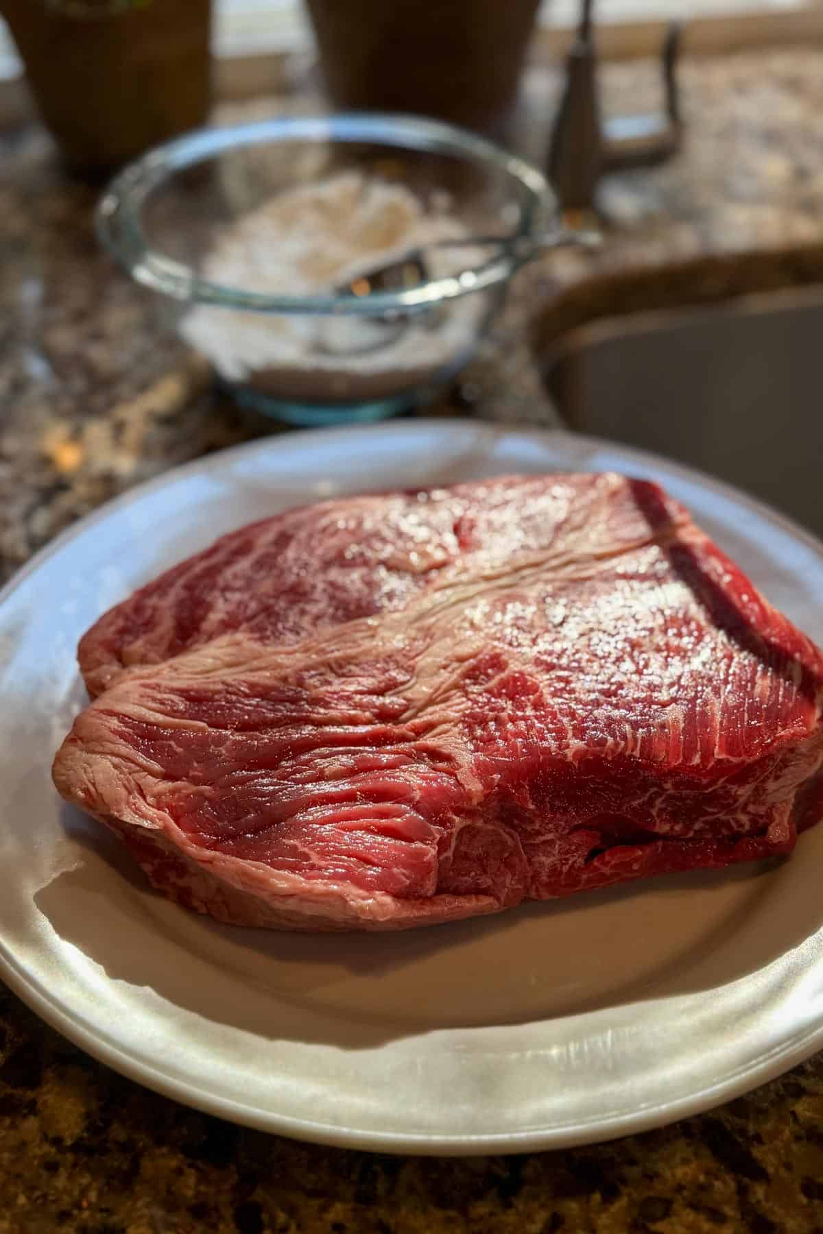 A close-up of a raw beef chuck roast on a white plate, placed on a marble countertop. In the background, a mixing bowl with a whisk can be seen. Sunlight highlights the marbled texture of the meat.