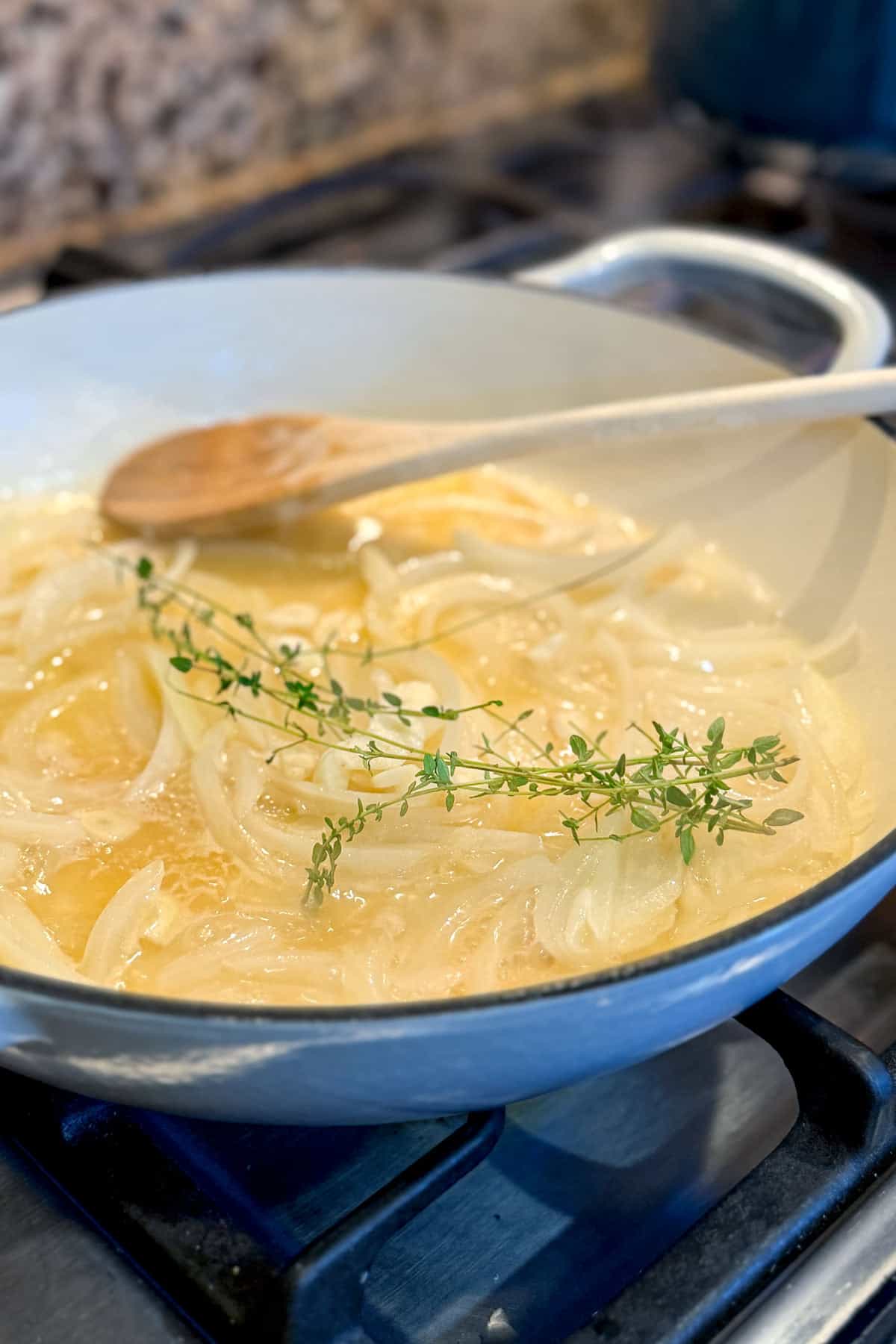 A pan on a stove contains sliced onions being sautéed in butter or oil, with a sprig of fresh thyme on top. A wooden spoon rests in the pan, and the stove background is slightly out of focus.