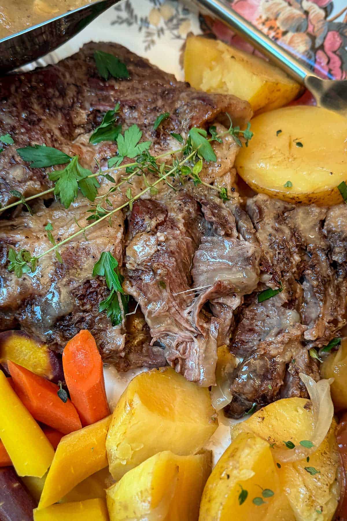 A close-up of a hearty meal featuring slices of roasted meat garnished with fresh herbs. Accompanying the meat are golden-brown potato wedges and vibrant slices of carrots.