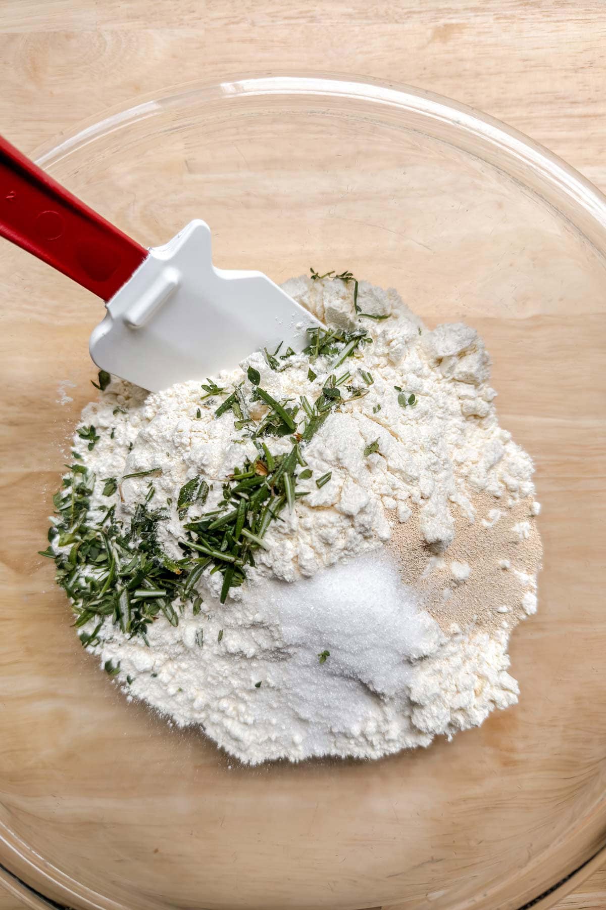 Dry ingredients of flour, salt, rosemary, and yeast in a glass bowl.