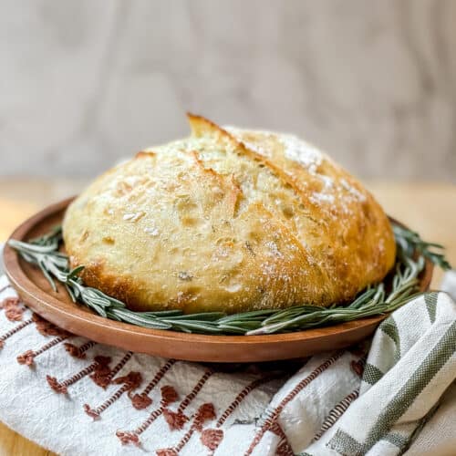 Loaf of freshly baked rosemary no knead bread with a ring of fresh rosemary on a wooden tray.
