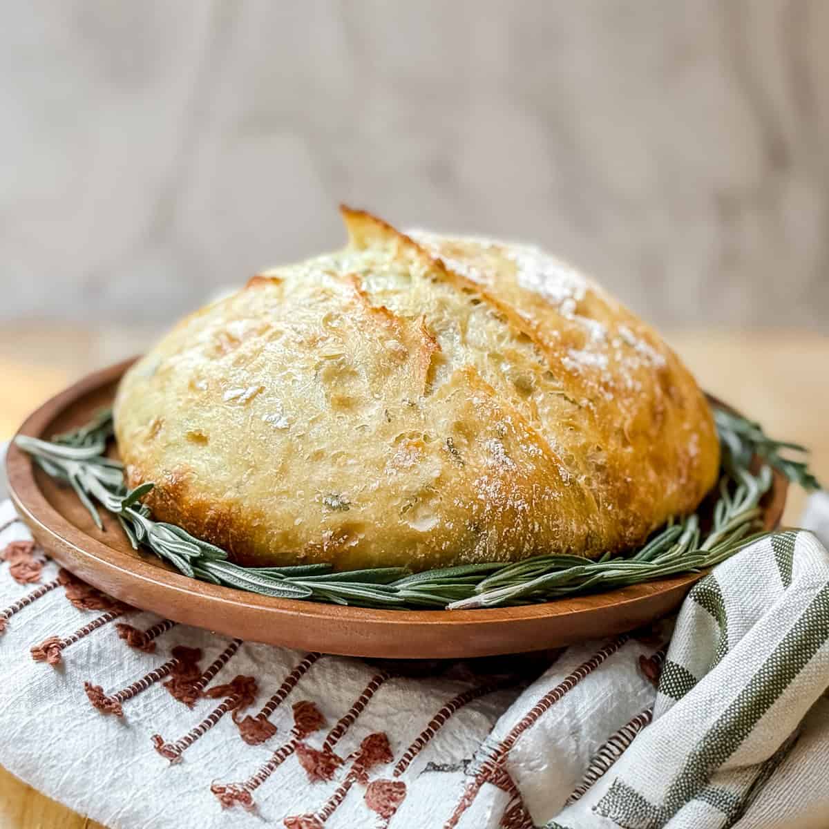 Loaf of freshly baked rosemary no knead bread with a ring of fresh rosemary on a wooden tray.