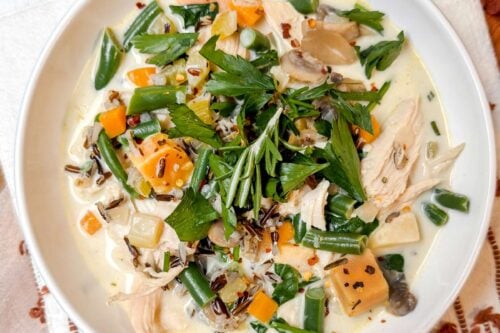 A white bowl filled with a creamy turkey wild rice soup. Garnished with fresh herbs, the bowl is placed on a wooden board with a white and brown embroidered cloth.