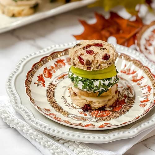 A stack of turkey tea sandwiches on cranberry bread with green apple slices and cream cheese, garnished with herbs. A teacup with red tea sits beside it, creating a cozy setting complete with autumn leaves on a white tablecloth.