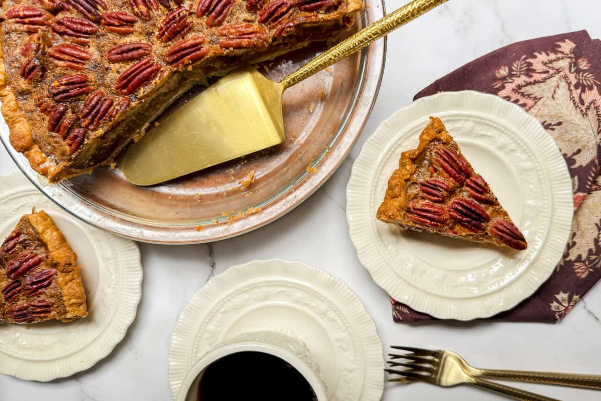 A pecan pie, a testament to a perfect recipe, has slices cut out and placed on white plates. A golden pie server rests in the dish. Nearby, a decorative napkin accompanies a cup of black coffee and a fork. The whole scene is elegantly set on a white marble surface.