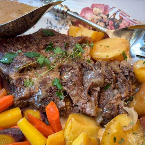 A delicious pot roast on a decorative plate, surrounded by roasted carrots and potatoes. A metal gravy boat with sauce and a serving fork are beside the dish. The roast is garnished with fresh parsley.