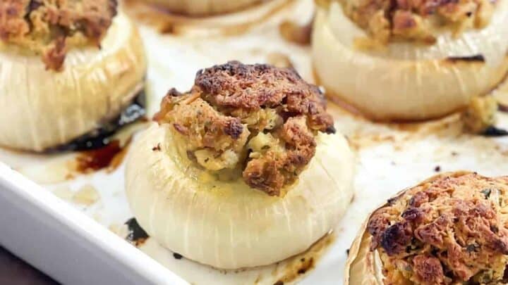 Five roasted onions stuffed with a savory breadcrumb mixture are arranged in a white baking dish. The onions are golden and slightly caramelized on top, sitting on a wooden surface.