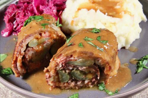 A plate featuring two pieces of rouladen topped with gravy, served alongside mashed potatoes with more gravy and a portion of red cabbage. A fork is seen in the background on the tablecloth.