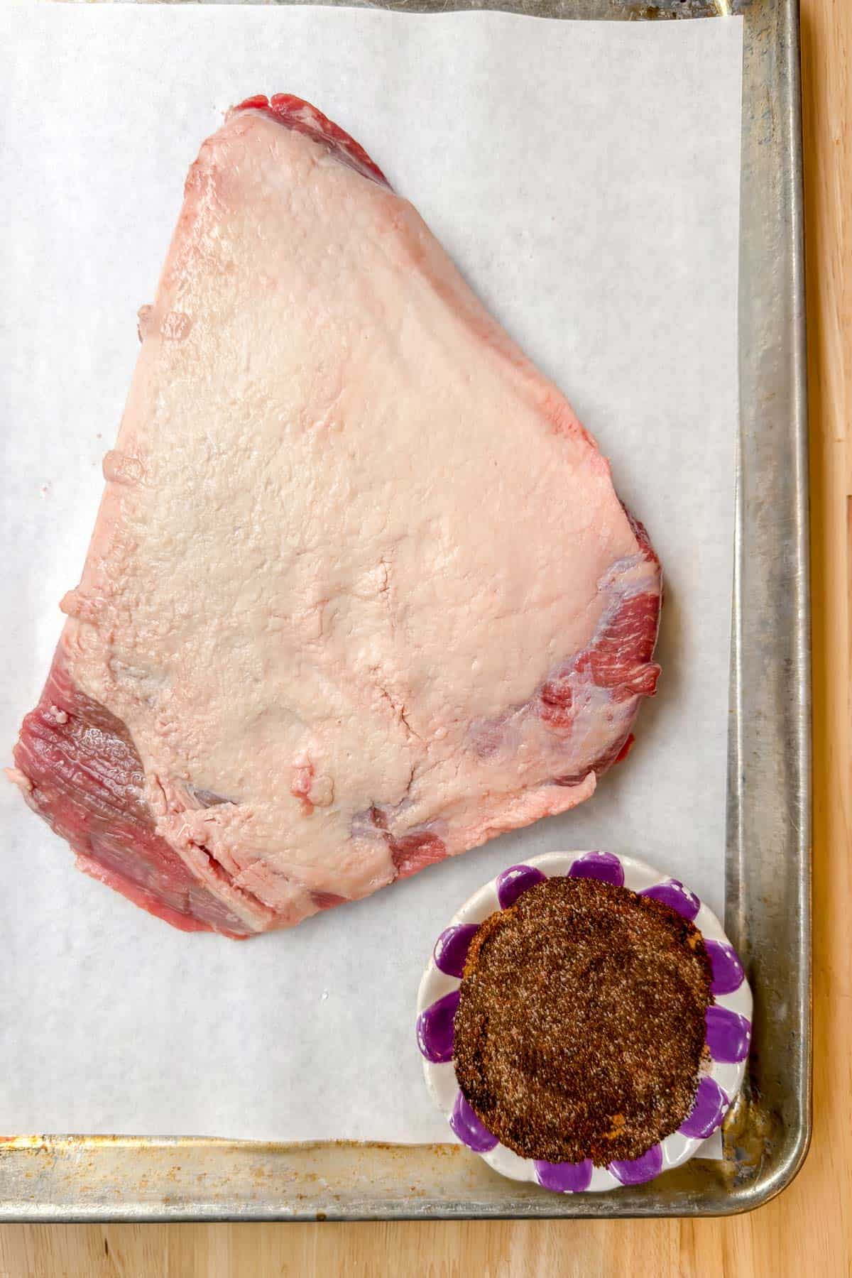 A raw beef brisket on a parchment-lined baking sheet, poised for transformation into a savory delight, rests beside a small dish of aromatic brown spices—an essential step in this flavorful brisket recipe.