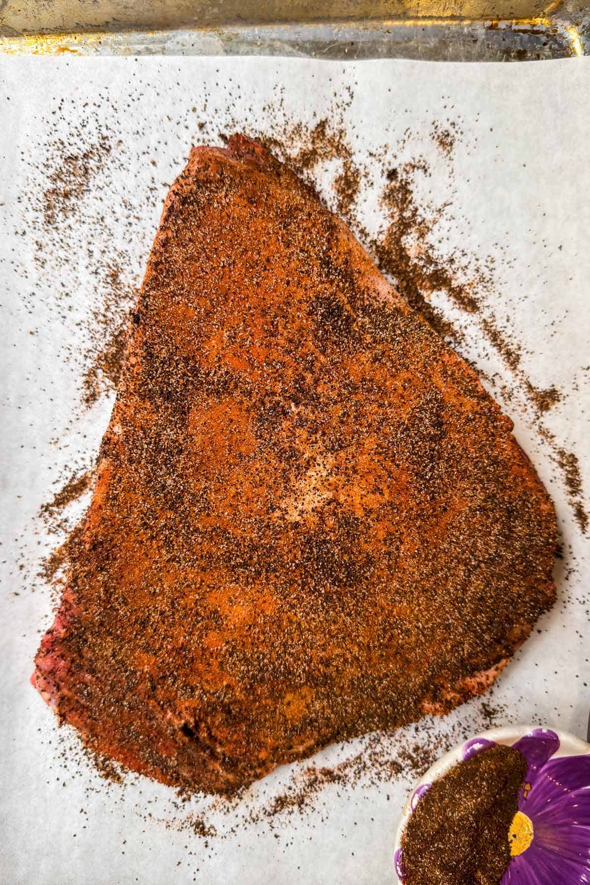 A raw piece of beef brisket, soon to star in a delectable brisket recipe, is covered in a dark spice rub and sits on white parchment paper. Nearby, a purple bowl holds extra spice mix. The coarse rub contrasts sharply against the light background, hinting at flavors to come.