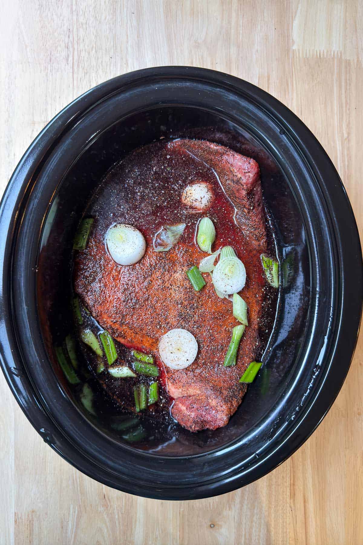 A slow cooker brimming with a brisket recipe showcases raw seasoned beef roast, chopped green onions, and petite whole onions submerged in broth, all set on a wooden table.