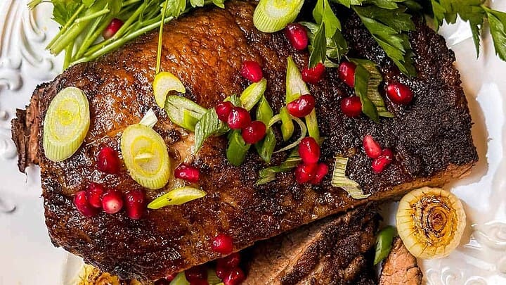 Slices of roasted brisket, garnished with pomegranate seeds, green onions, and parsley on a white platter.