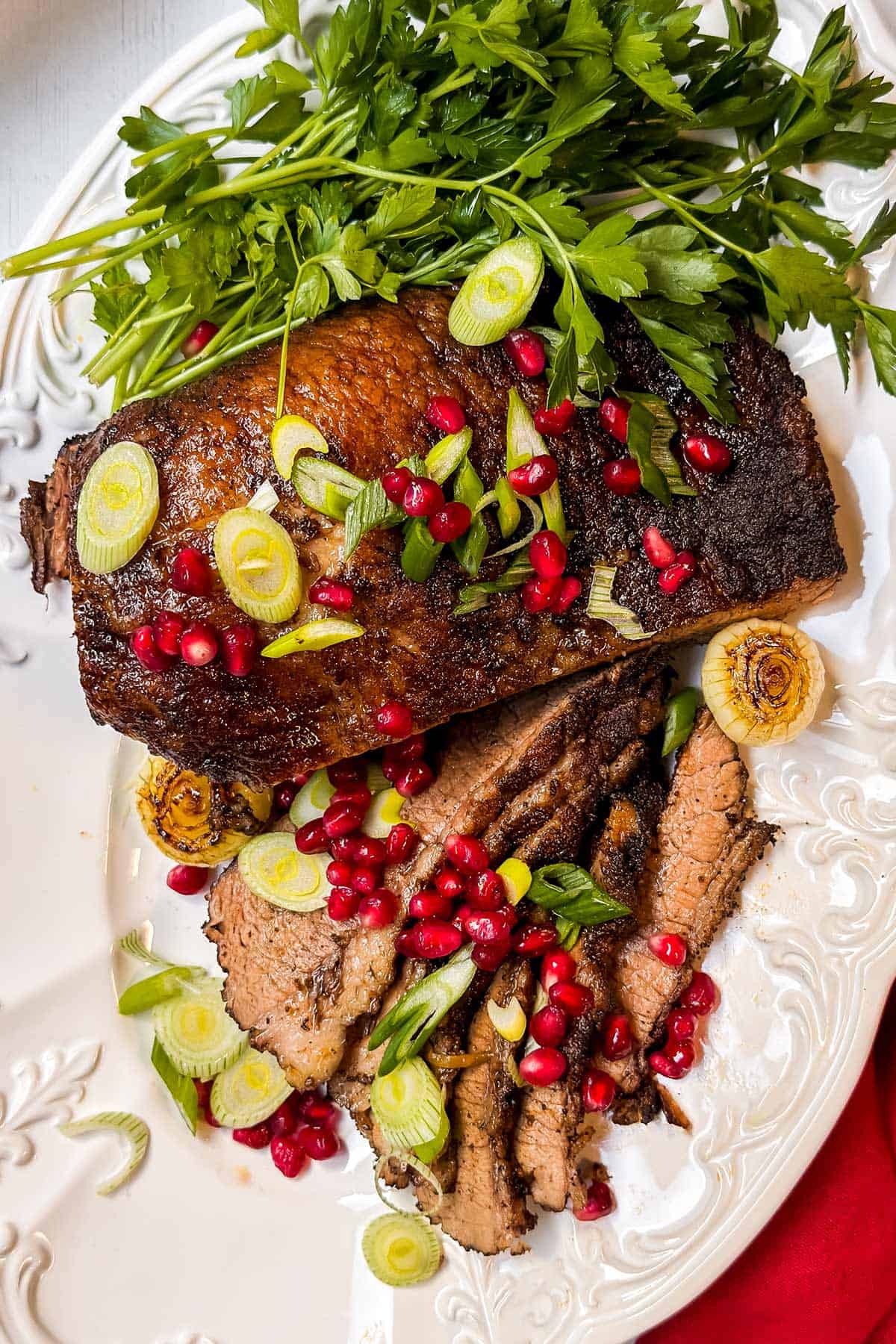 Slices of roasted brisket, garnished with pomegranate seeds, green onions, and parsley on a white platter.