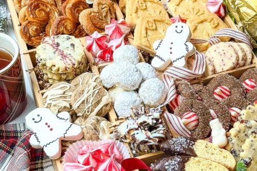 A festive wooden tray filled with a variety of holiday cookies, including gingerbread men, powdered sugar cookies, biscotti, and peppermint swirls. Surrounded by a plaid cloth and winter greenery, the scene is cozy and inviting with a red ribbon accent.