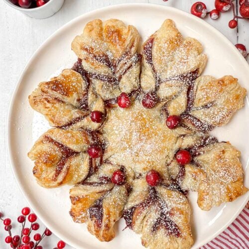 A festive puff pastry shaped like a star, dusted with powdered sugar and topped with cranberries, sits on a round plate. Surrounding the plate are red berries and a small bowl of cranberries, evoking a Christmas tree scene. A striped cloth napkin rests at the bottom.