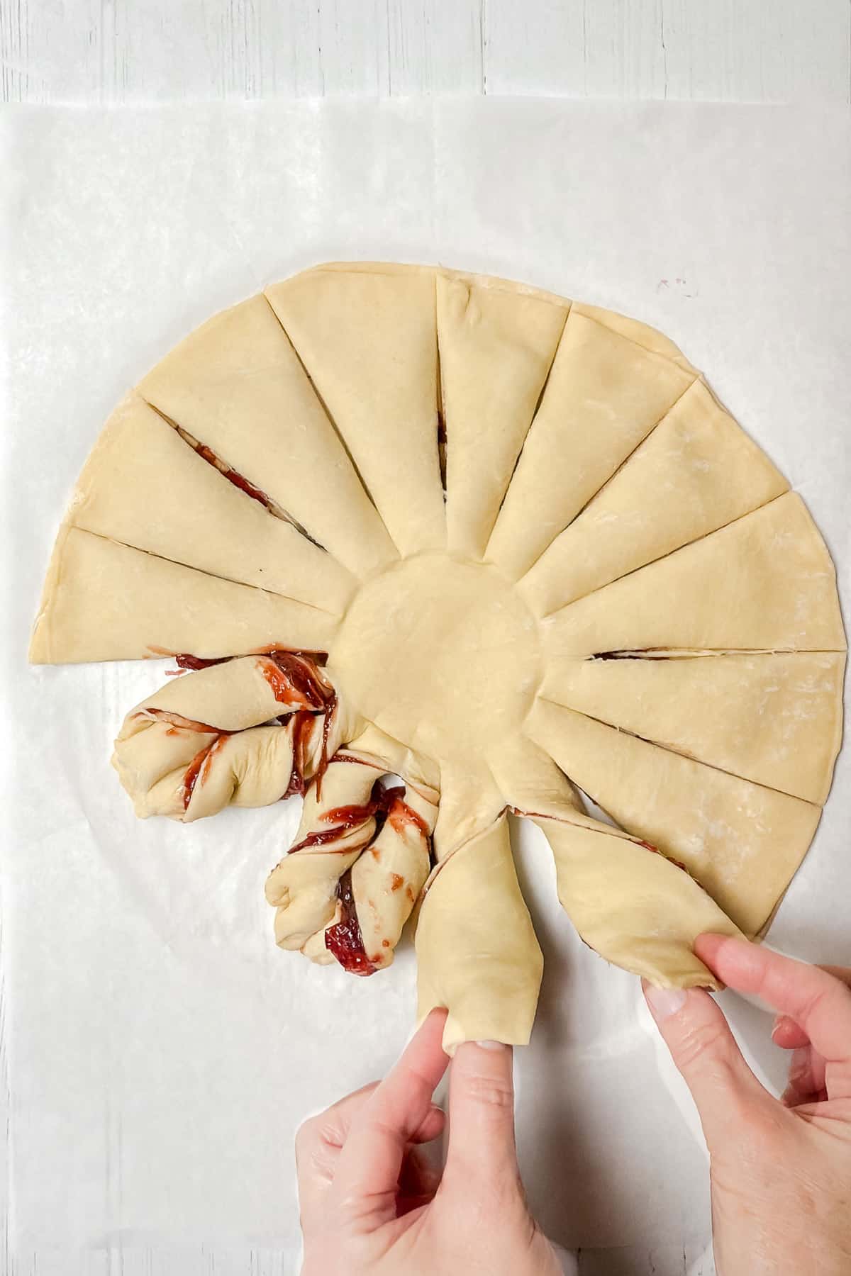 Hands are twisting sections of a circular puff pastry dough with cranberry filling, laid on parchment paper. The dough is cut into triangular segments, resembling a sunburst pattern.