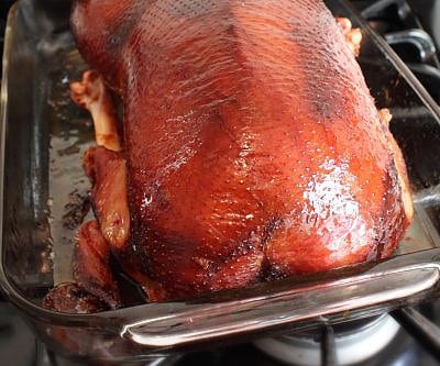 A whole roasted duck with crispy skin is placed in a glass baking dish on a stove. The duck is richly browned, and its legs are visible, resting on the edge of the dish.