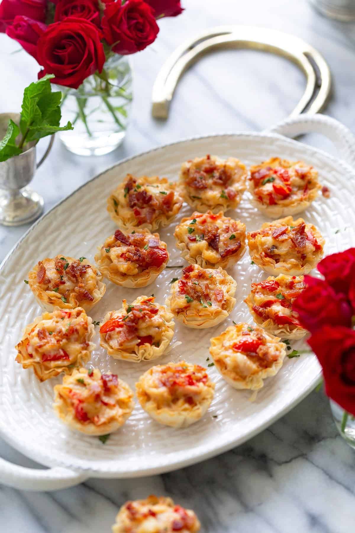 An oval platter with mini tarts topped with diced tomatoes, herbs, and melted cheese. The platter is set on a marble surface, surrounded by red roses in silver vases.