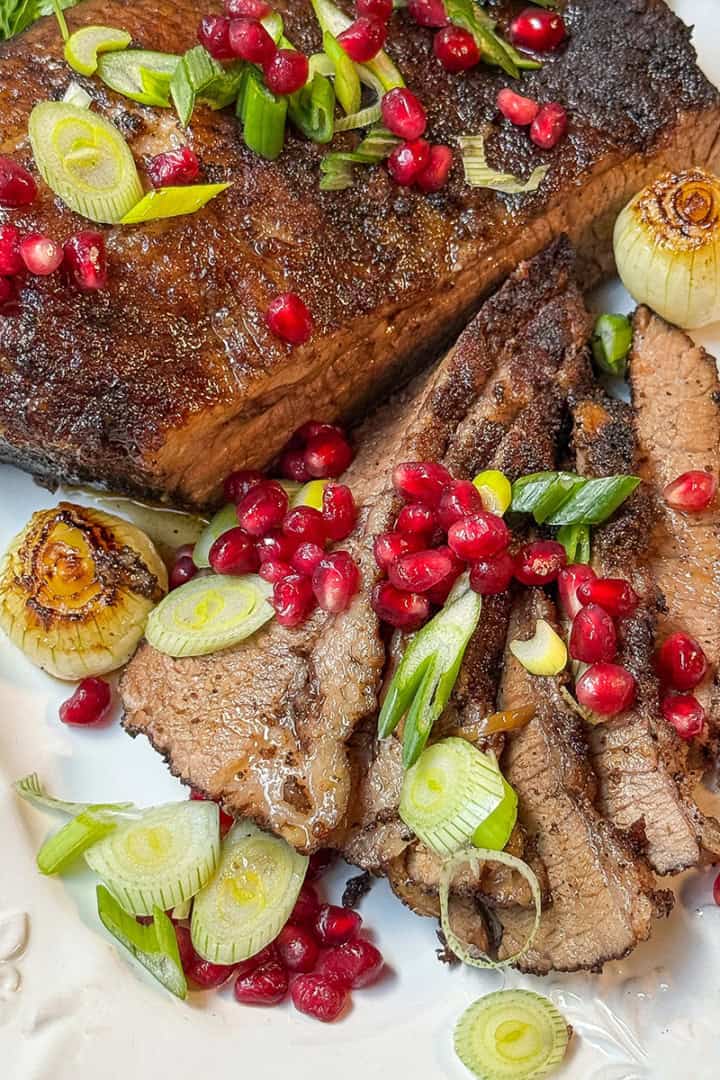 Square image of sliced slow cooker beef brisket on a white serving platter with pomegranate seeds.