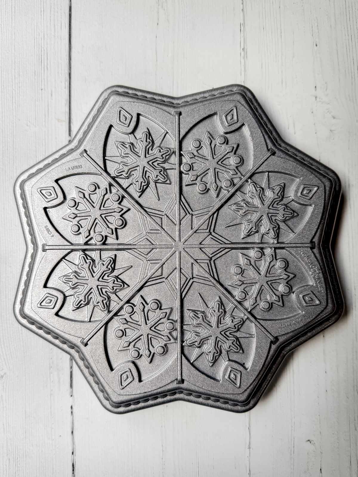 A metal baking pan with a winter-themed design features sections for crafting six snowflake-shaped cakes or delightful shortbread cookies. The pan rests elegantly on a white wooden surface.