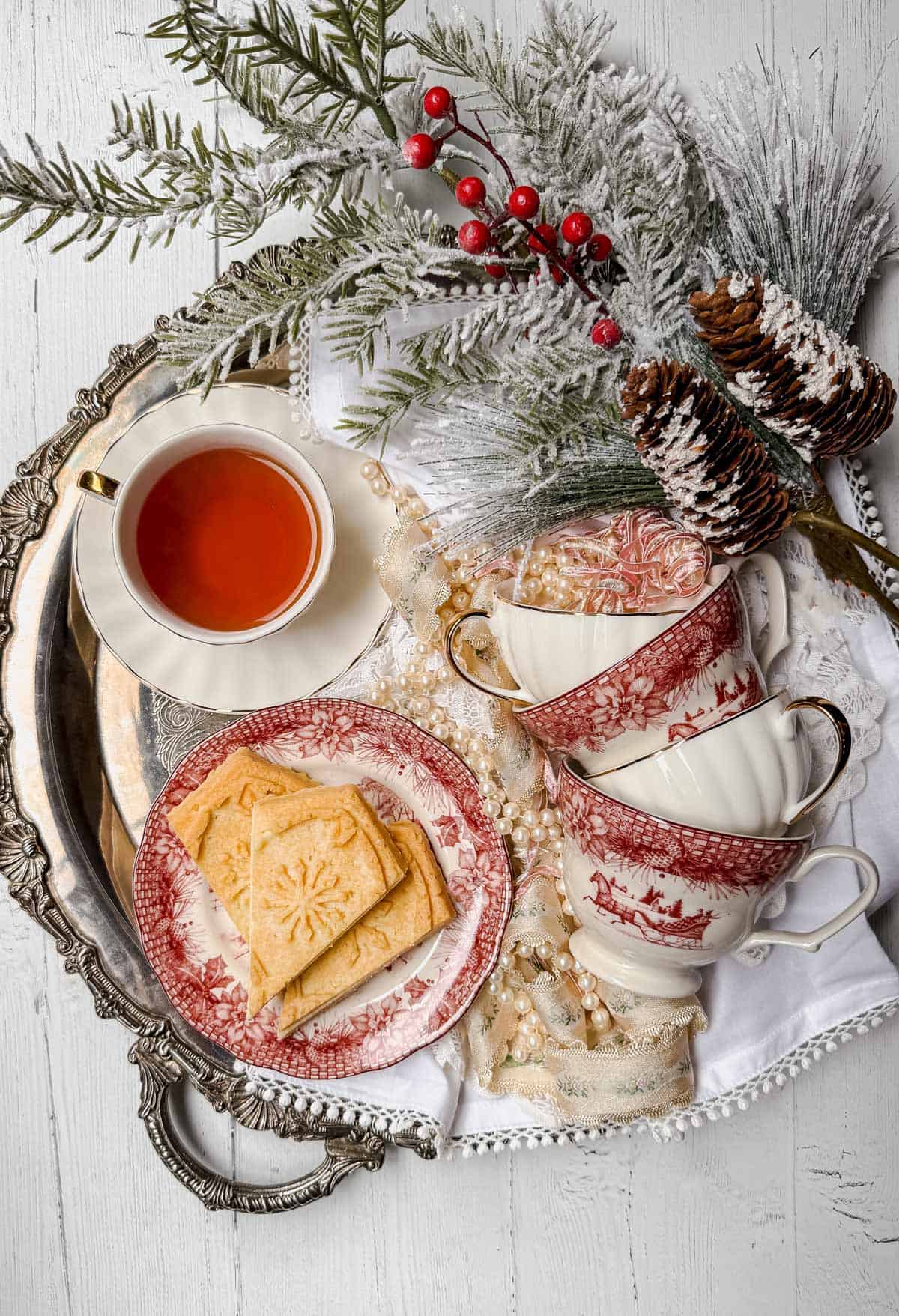 A decorative tray holds a cup of tea, stacked red and white teacups, a plate of shortbread cookies with festive designs, snow-dusted pinecones, red berries, and evergreen branches, creating a cozy, winter-themed setting on a white wooden surface.