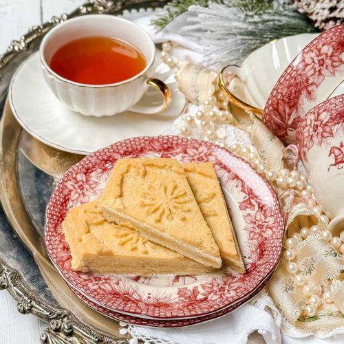 A silver tray holds a steaming cup of tea and delectable shortbread cookies. The plate, adorned with red and white patterns, adds charm to the scene. Festive greenery and vibrant red berries enhance the background, evoking a cozy holiday theme.