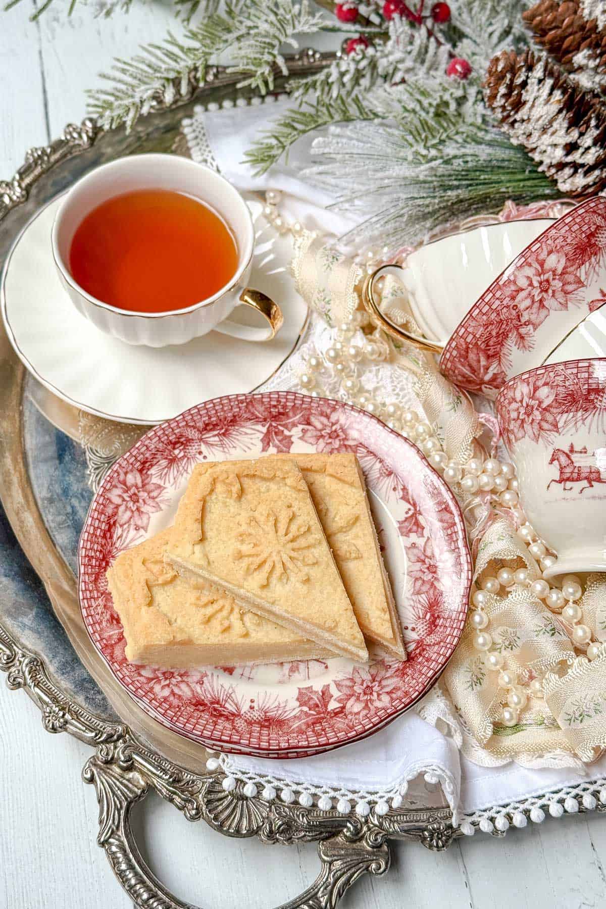 A festive scene features tea in an elegant cup and two scrumptious shortbread cookies on a decorative plate. The tray is adorned with winter-themed details, including greenery, berries, and pinecones, while extra teacups elegantly await nearby.