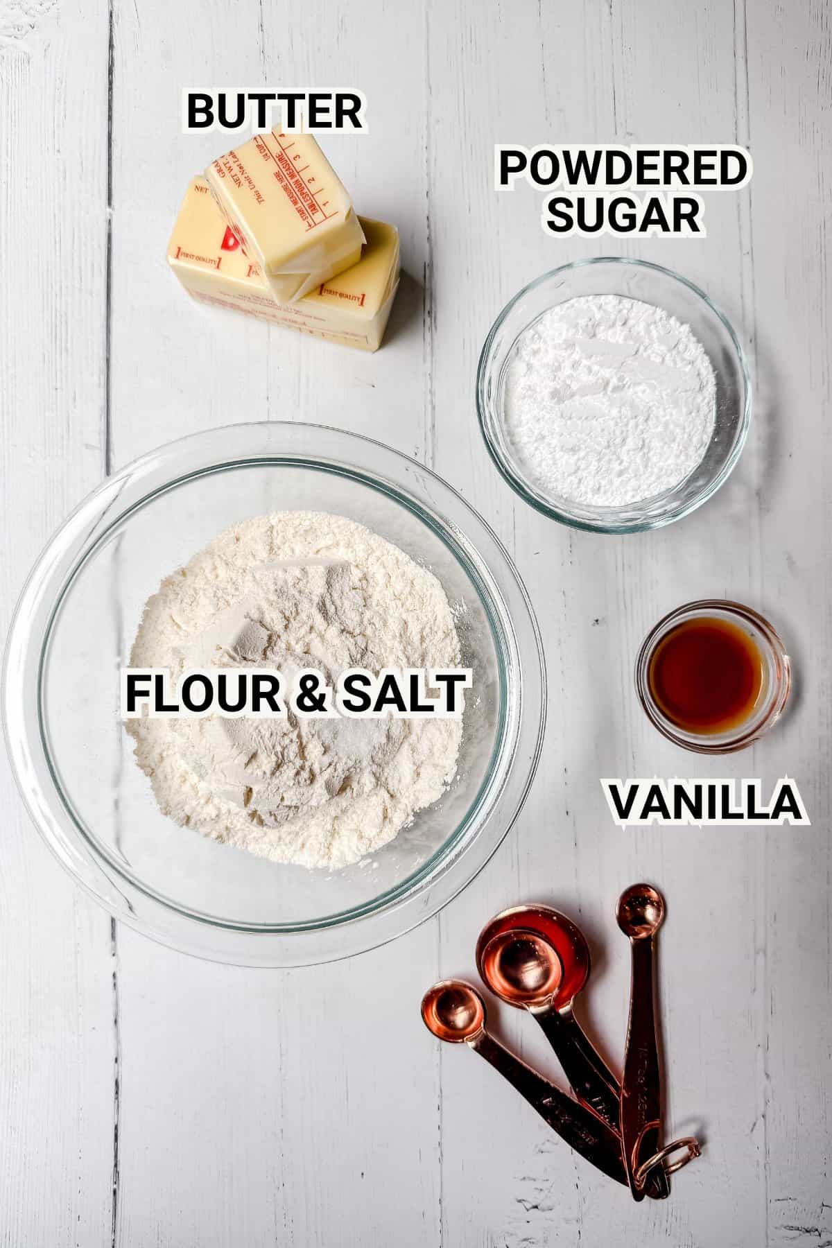 Top-down view of shortbread cookie ingredients on a white wooden surface. Includes labeled bowls of flour & salt, powdered sugar, a stick of butter, a small cup of vanilla extract, and copper measuring spoons.