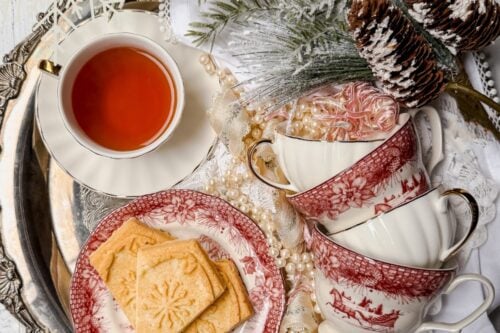 A festive tray features a cup of tea, stacked red and white china teacups, and snowflake-embossed shortbread cookies. Adorned with pine cones, branches, and snowflakes, the tray showcases a "Snowflake Shortbread Cookies" banner at the top.