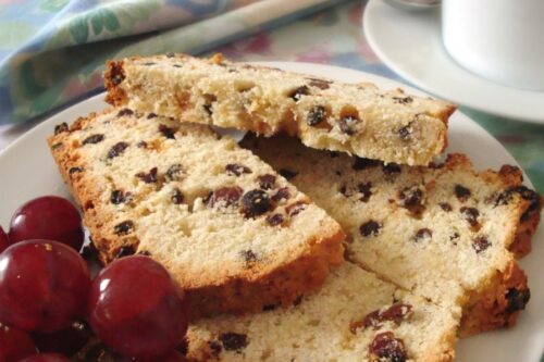 Slices of raisin bread on a white plate with red grapes. A colorful cloth napkin and a cup of coffee are in the background.
