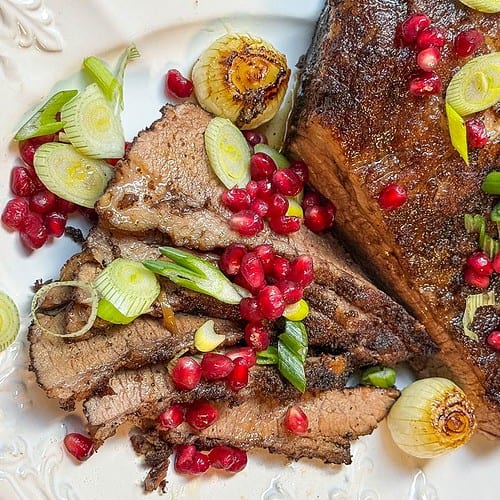 Sliced roasted brisket garnished with pomegranate seeds and green onions graces a white plate. Whole caramelized onions and fresh parsley accompany this savory brisket recipe, creating a stunning presentation.