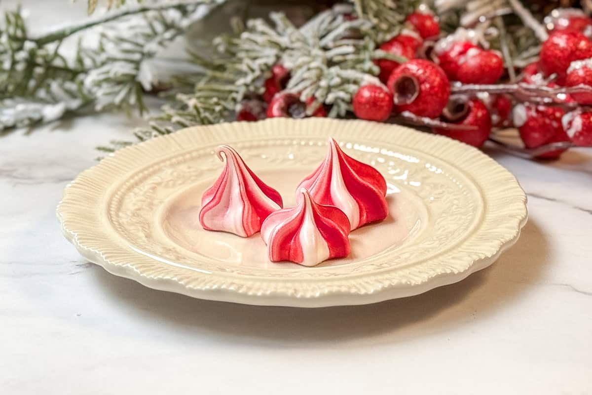 Three red and white swirled meringue cookies are artfully arranged on an ornate, off-white plate. Festive greenery and red berries in the background enhance the holiday touch.