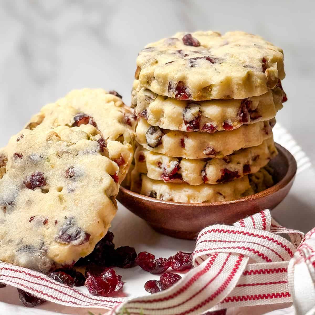Stack of Cranberry Pistachio Shortbread Cookies on a white stand with red and white striped ribbon and dried cranberries on the plate.