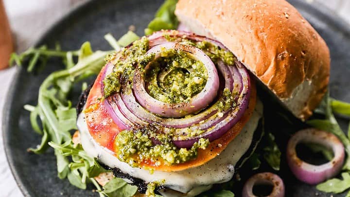 A gourmet veggie burger on a black plate, topped with grilled red onion, tomato, melted cheese, and pesto sauce, with fresh arugula. A partially open bun sits on the side, with a bowl of pesto in the background.
