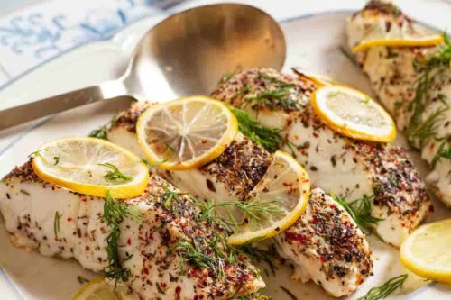 A plate of seasoned baked fish fillets, perfect for January 20-minute dinners, is garnished with lemon slices and fresh dill. A metal serving spoon sits beside it. The background features small dishes with seasoning and salt, while a red cloth napkin adds a pop of color in the foreground.