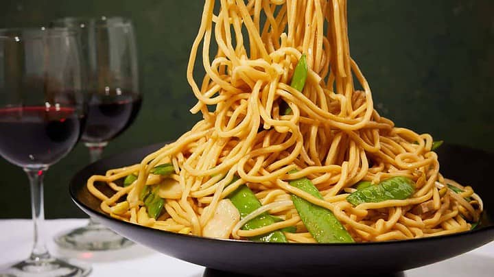 A bowl of stir-fried noodles with snap peas being served with tongs. Two glasses of red wine are in the background on a white tablecloth. The noodles are golden brown and garnished with vegetables.