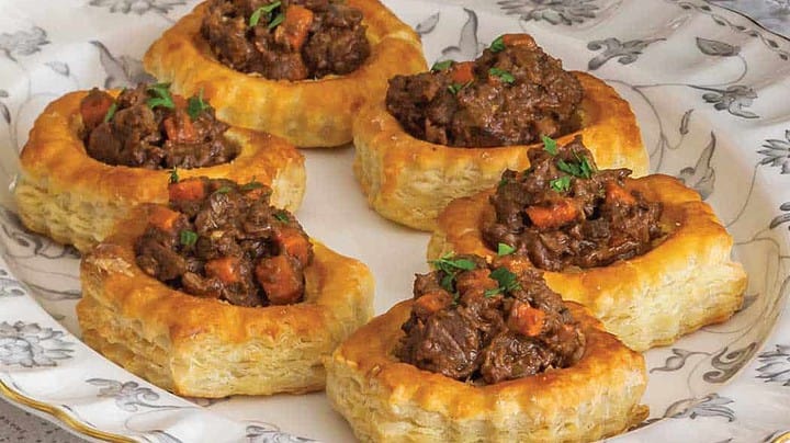 A plate of six savory puff pastry vol-au-vents filled with a hearty beef stew, garnished with chopped herbs, displayed on an elegant floral-patterned dish.