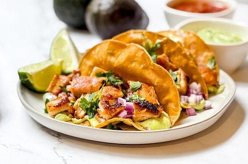 Three crispy chicken tacos filled with grilled goodness, garnished with cilantro and diced red onions, are placed on a white plate. Lime wedges sit on the side, accompanied by bowls of sauce and a blurred avocado in the background.