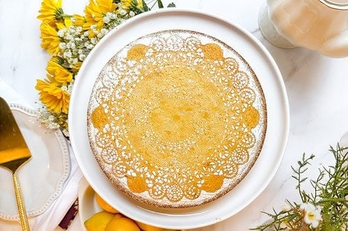 A round lemon cake with a decorative powdered sugar lace pattern on top rests elegantly on a white cake stand. Surrounded by fresh sunflowers and vibrant lemons, it exudes charm, while a golden cake server awaits nearby-a perfect setting to inspire your next lemon cake recipe.