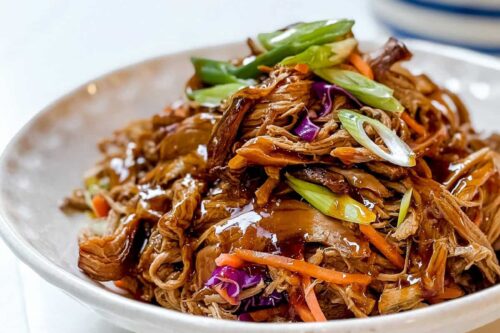 A dish of Asian shredded beef with a glossy sauce is garnished with sliced green onions and mixed vegetables like purple cabbage and carrots. The plate rests on a white surface, with a blurred round pot in the background.