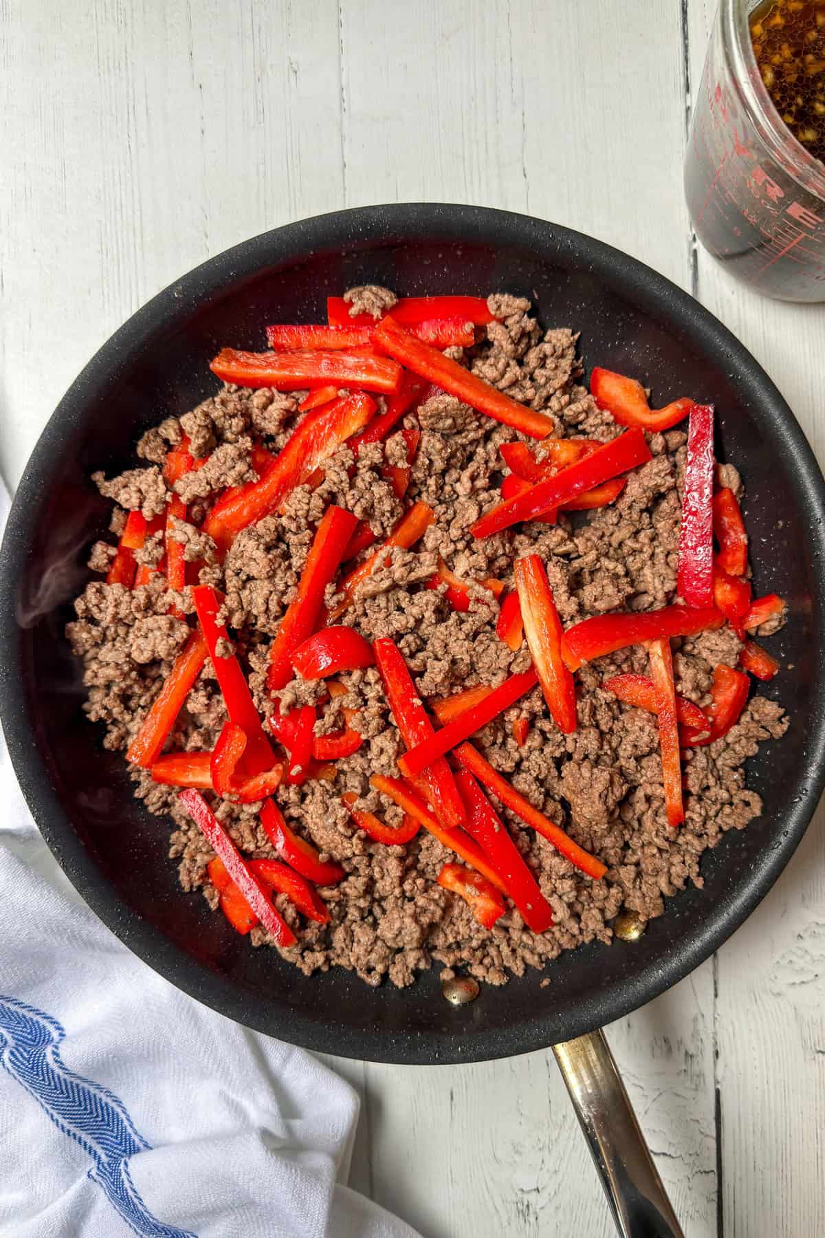 A black skillet containing cooked ground beef, sliced red bell peppers, and hints of Beef and Broccoli flavors sits on a white wooden surface. A measuring cup with liquid and a white towel are nearby.