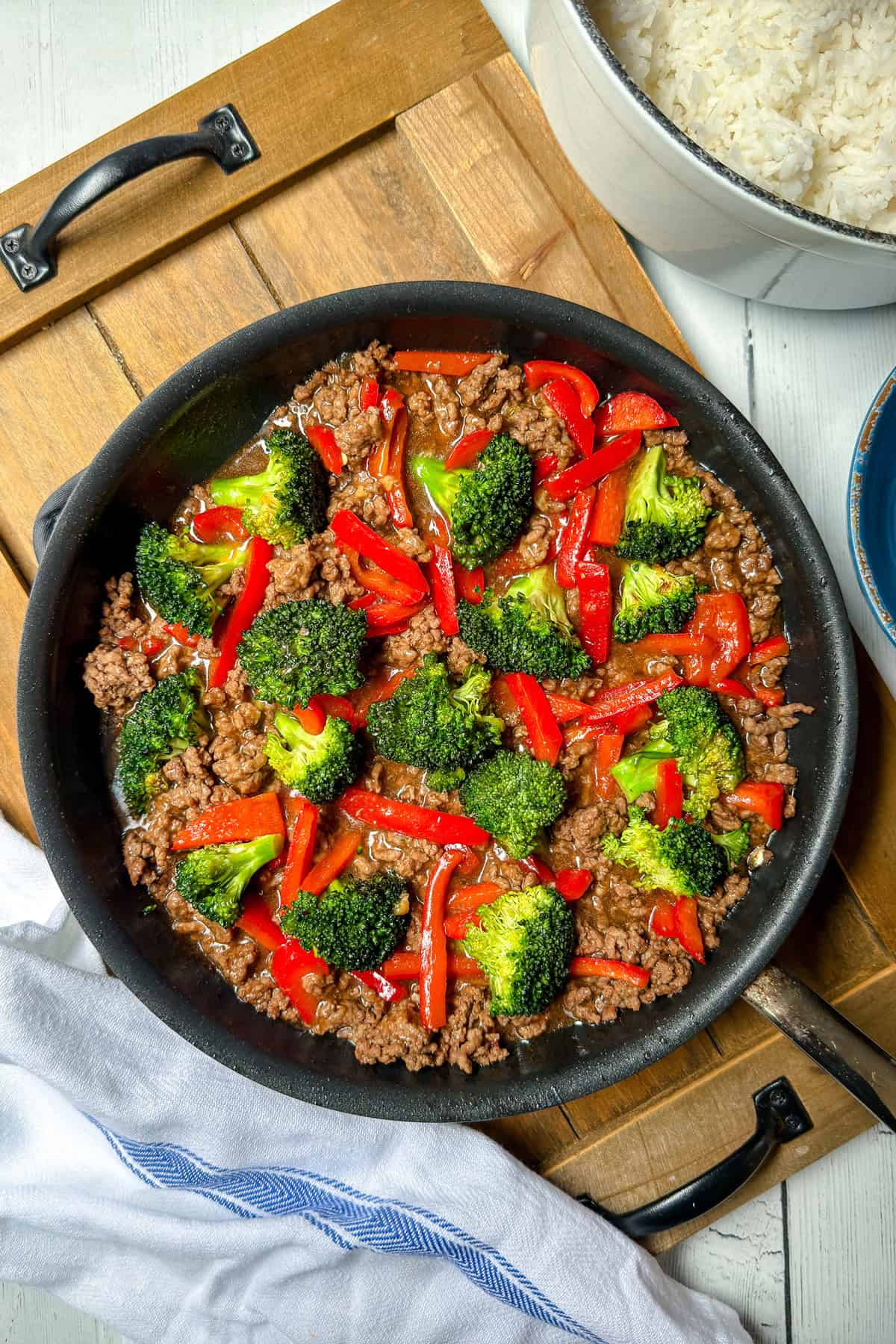 A skillet filled with Beef and Broccoli, ground beef, and red bell peppers in sauce, placed on a wooden board next to a pot of white rice.
