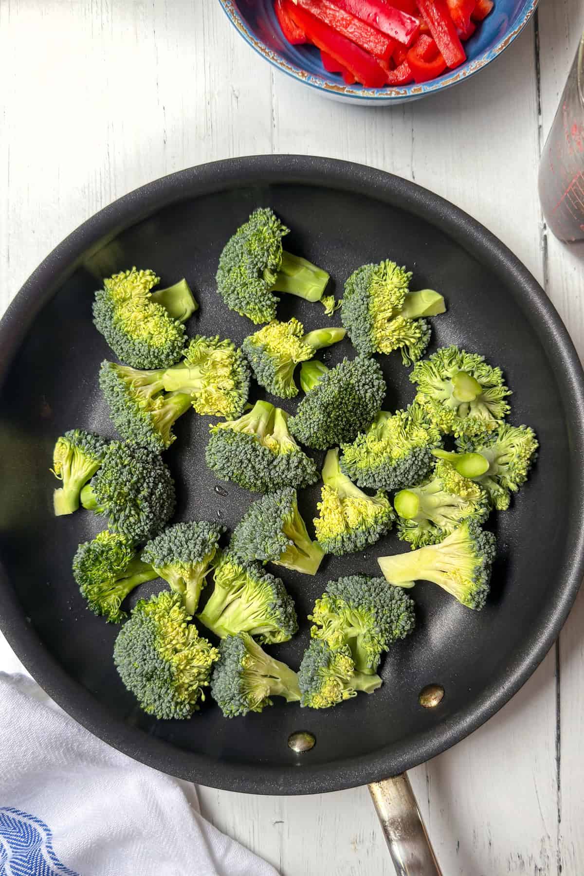 Broccoli florets are arranged in a single layer inside a black frying pan on a light wooden surface, ready to be mixed with beef for a classic Beef and Broccoli dish, with a bowl of sliced red bell peppers nearby.
