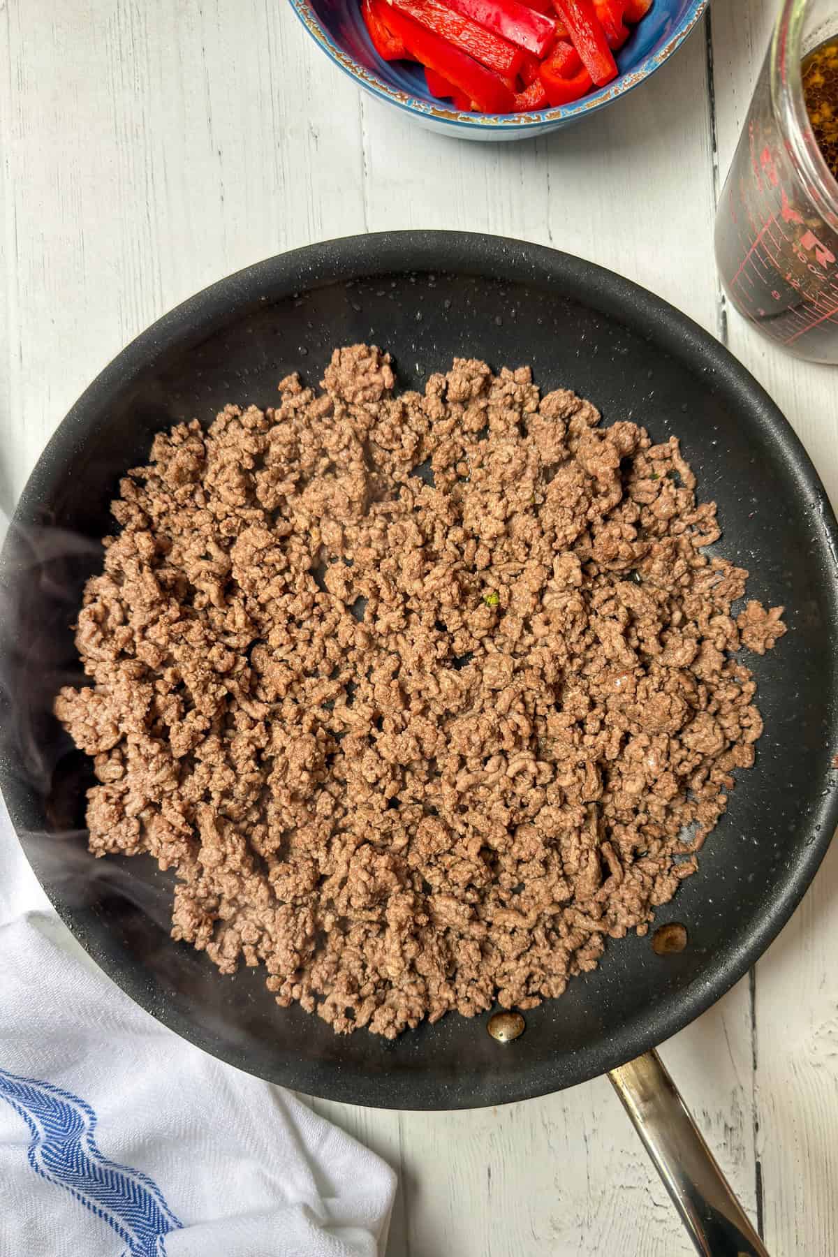 A frying pan filled with cooked ground beef, perfect for beef and broccoli recipes, sits on a white wooden surface, with a towel, a bowl of sliced red peppers, and a measuring cup nearby.