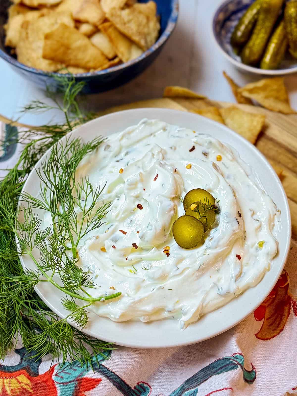 A plate of creamy dill pickle dip topped with three sliced pickles and fresh dill, surrounded by pita chips, more fresh dill, and a bowl of pickles in the background.