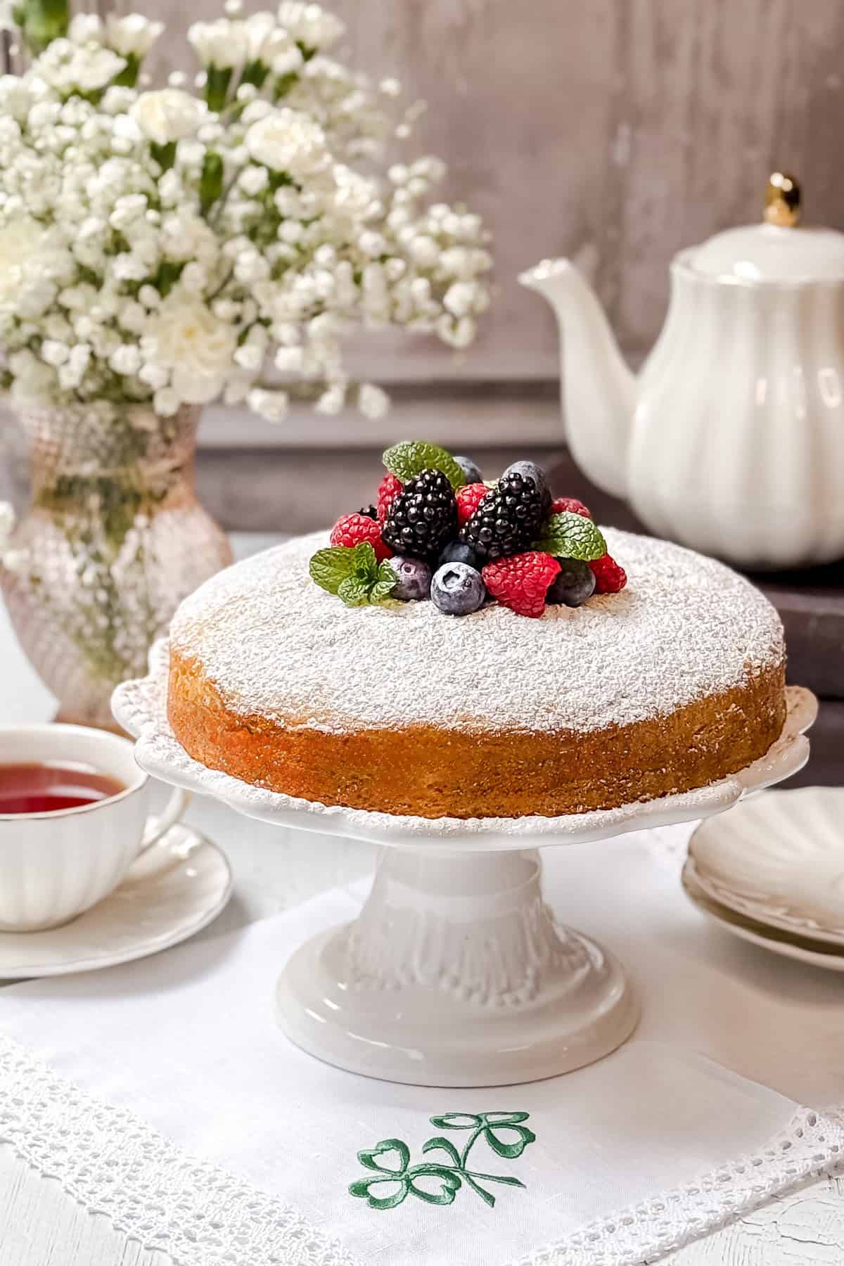 An Irish Tea Cake dusted with powdered sugar, topped with mixed berries, sits elegantly on a white cake stand. In the background, a teapot, teacup with tea, and a vase with white flowers rest on an embroidered white cloth.